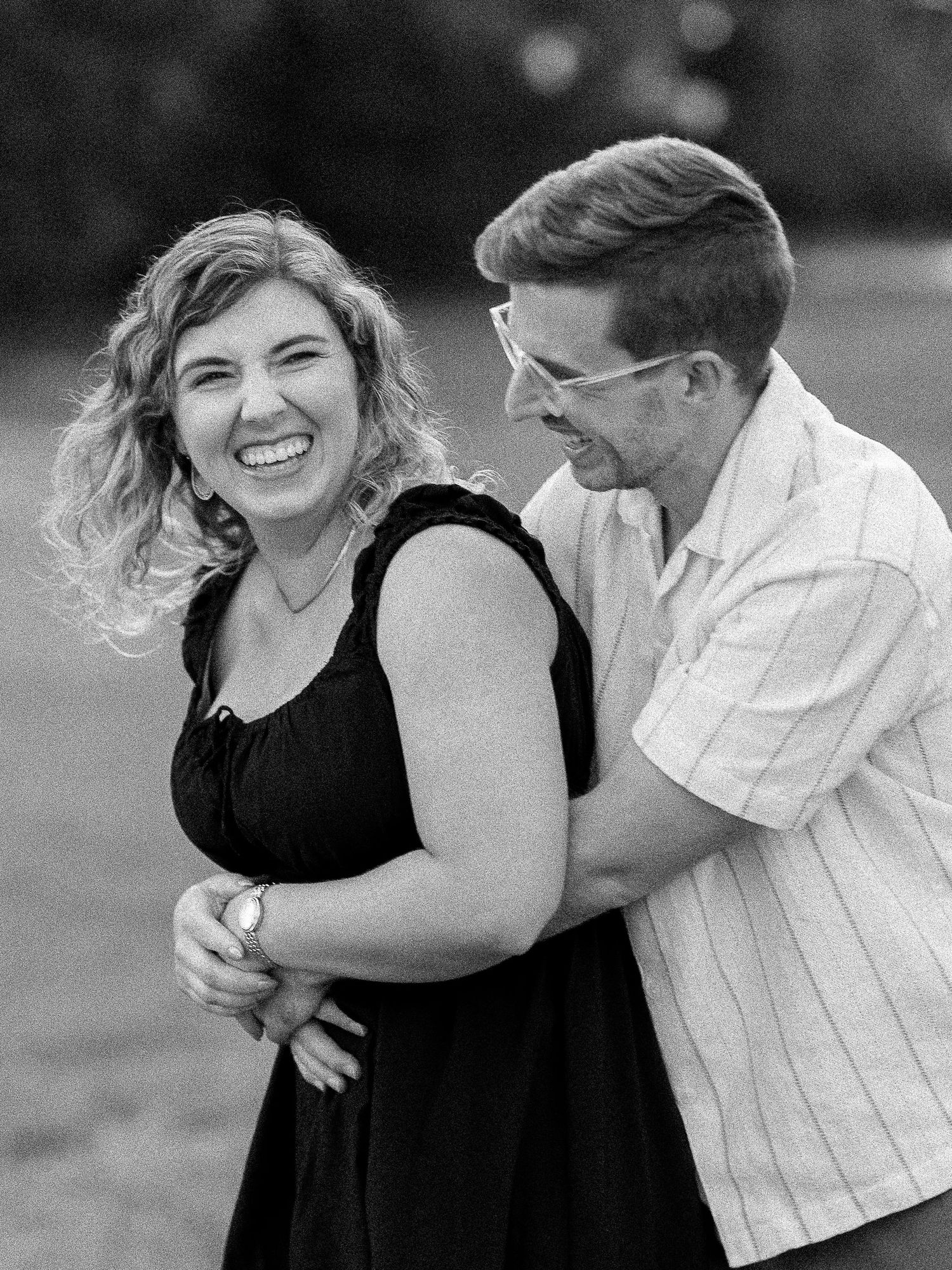 Black and white photo of a laughing couple embracing outdoors, woman in black dress, man in striped shirt wearing glasses.