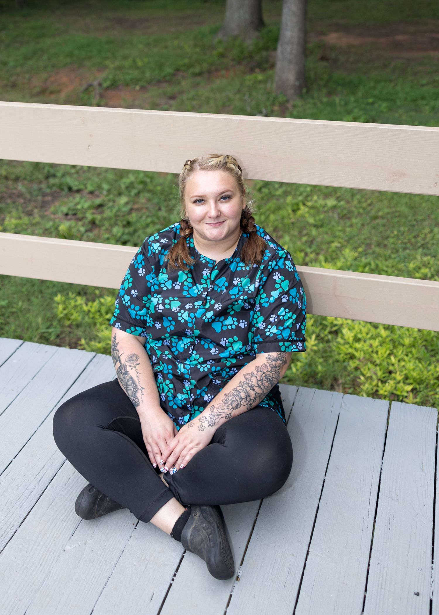 Someone wearing a blue floral print top and black pants sits cross-legged on a gray wooden deck outdoors.