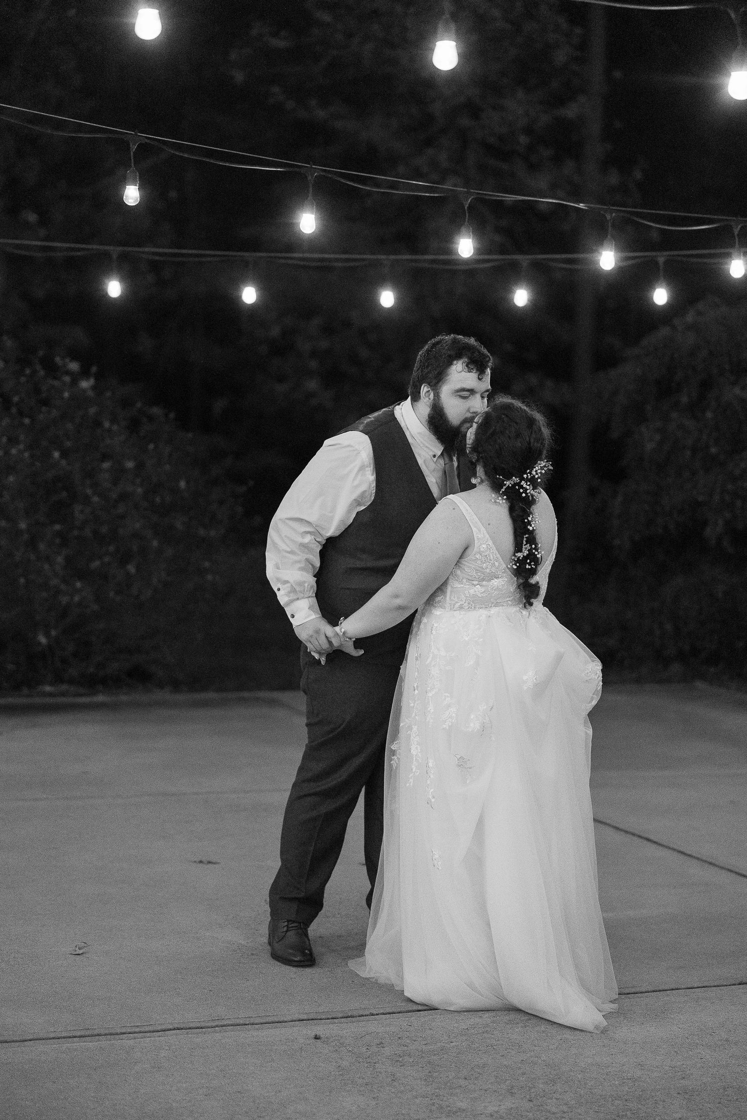 Black and white photo of a bride and groom sharing their first dance outdoors under string lights at night.