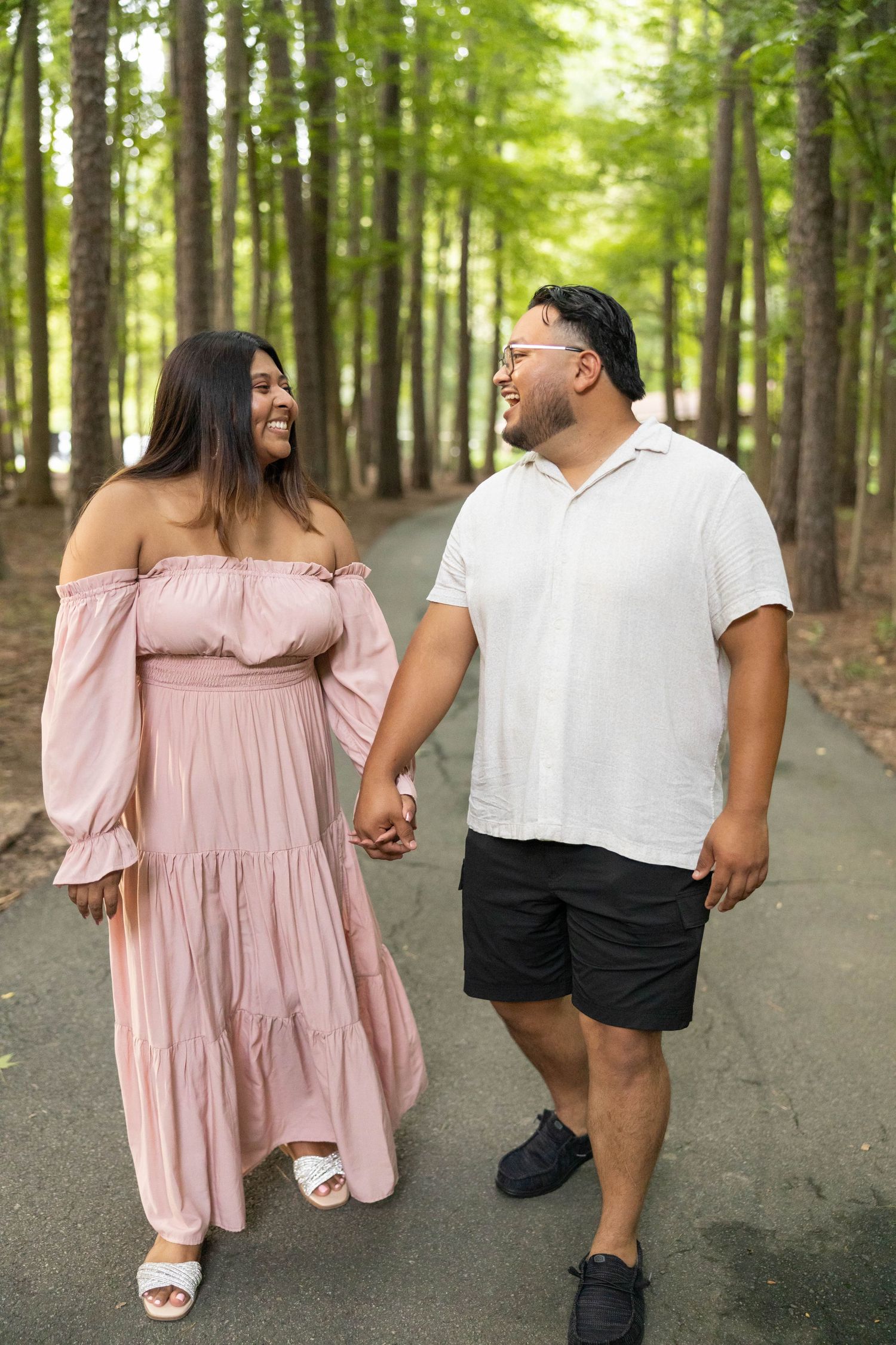 A couple walks hand in hand down a tree-lined path, with her wearing a pink dress and him in white shirt and black shorts.