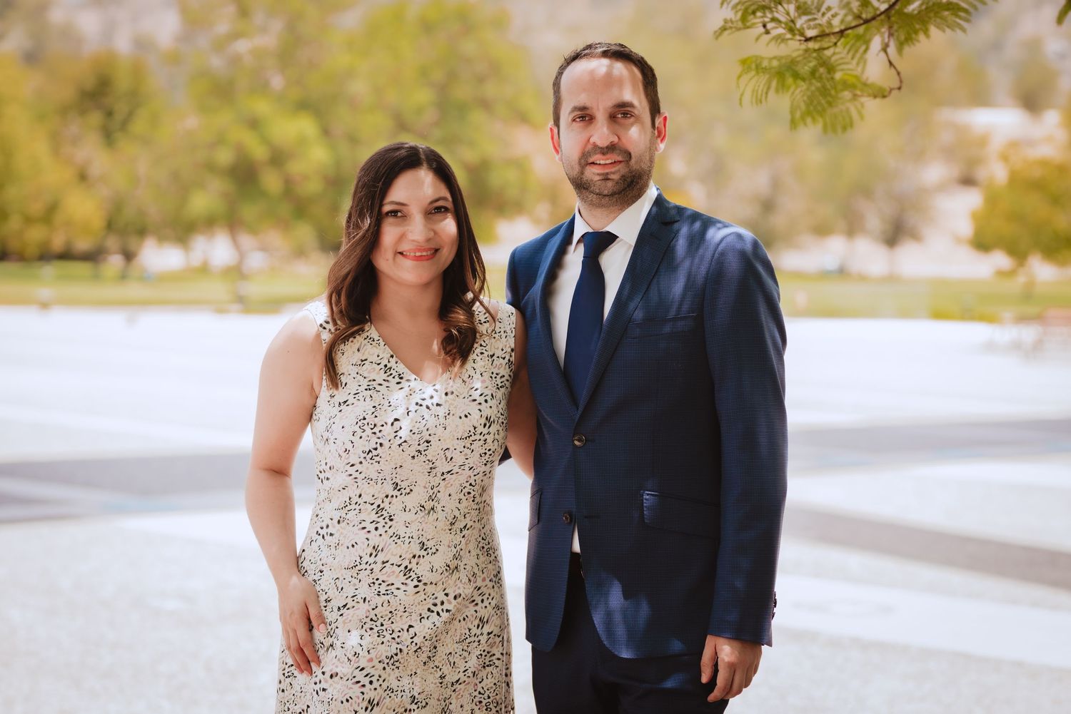 A couple in formalwear stands together outdoors against a blurred nature background.