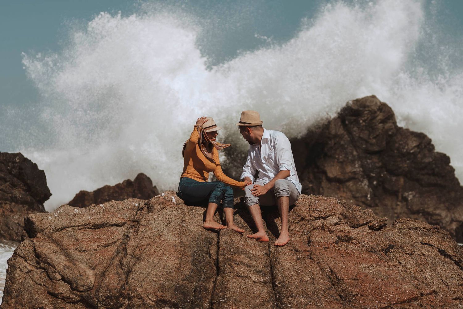 A couple sits together on rocky coastal cliffs with dramatic ocean waves crashing behind them.