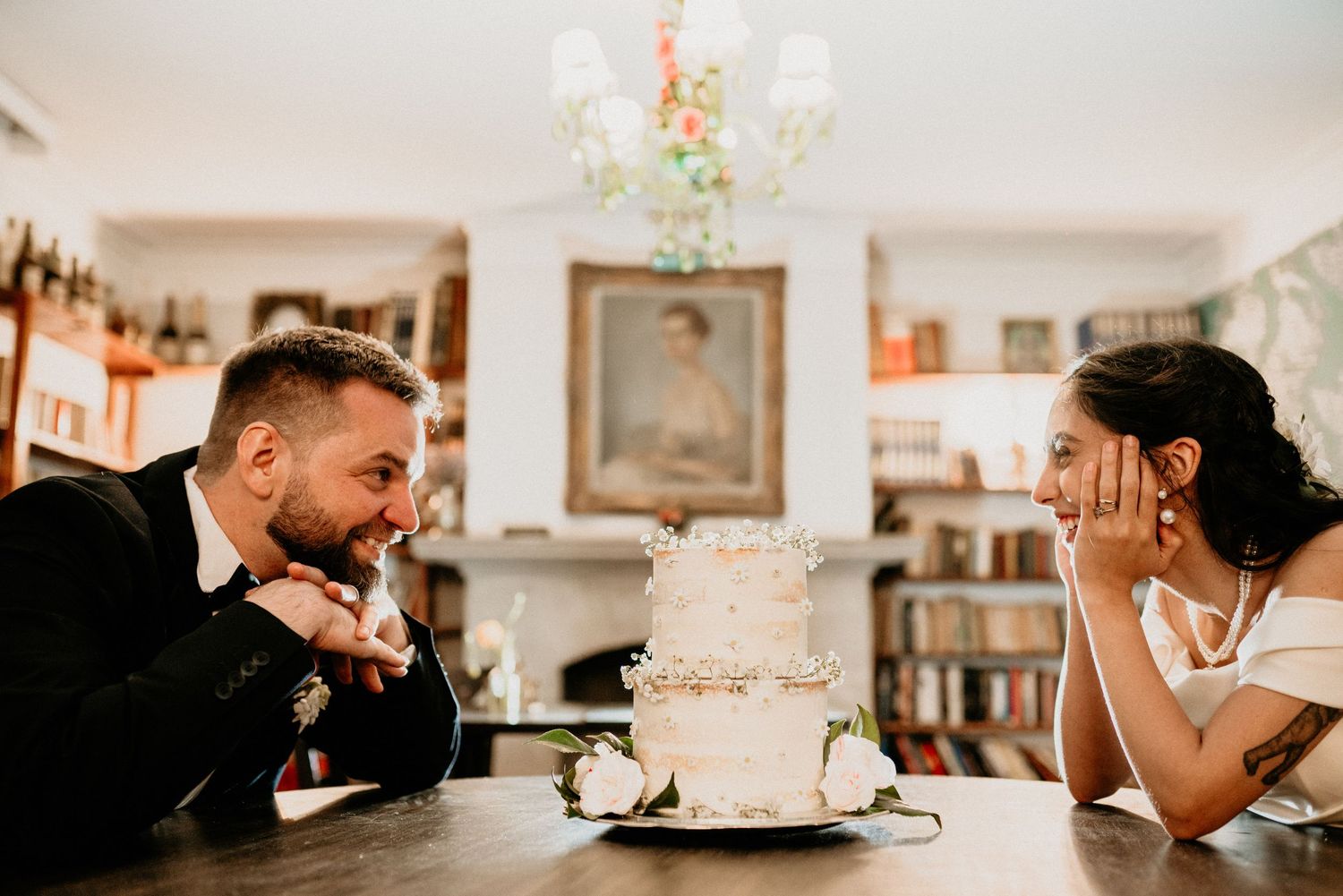 Couple sits at table with elegant white tiered wedding cake and chandelier in background during intimate celebration.