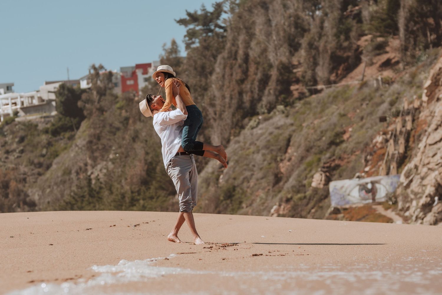 People walking along scenic beach with rocky cliffs and blue sky in background.