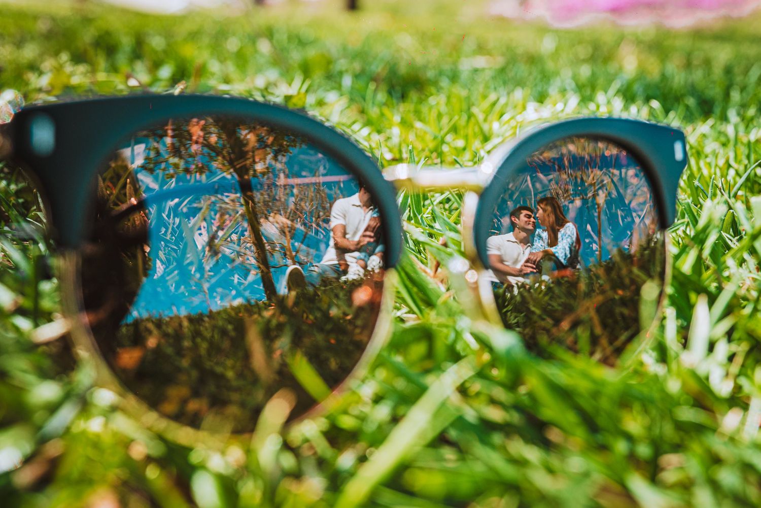 Creative reflection of a couple through sunglasses lens with blue sky and trees in background.