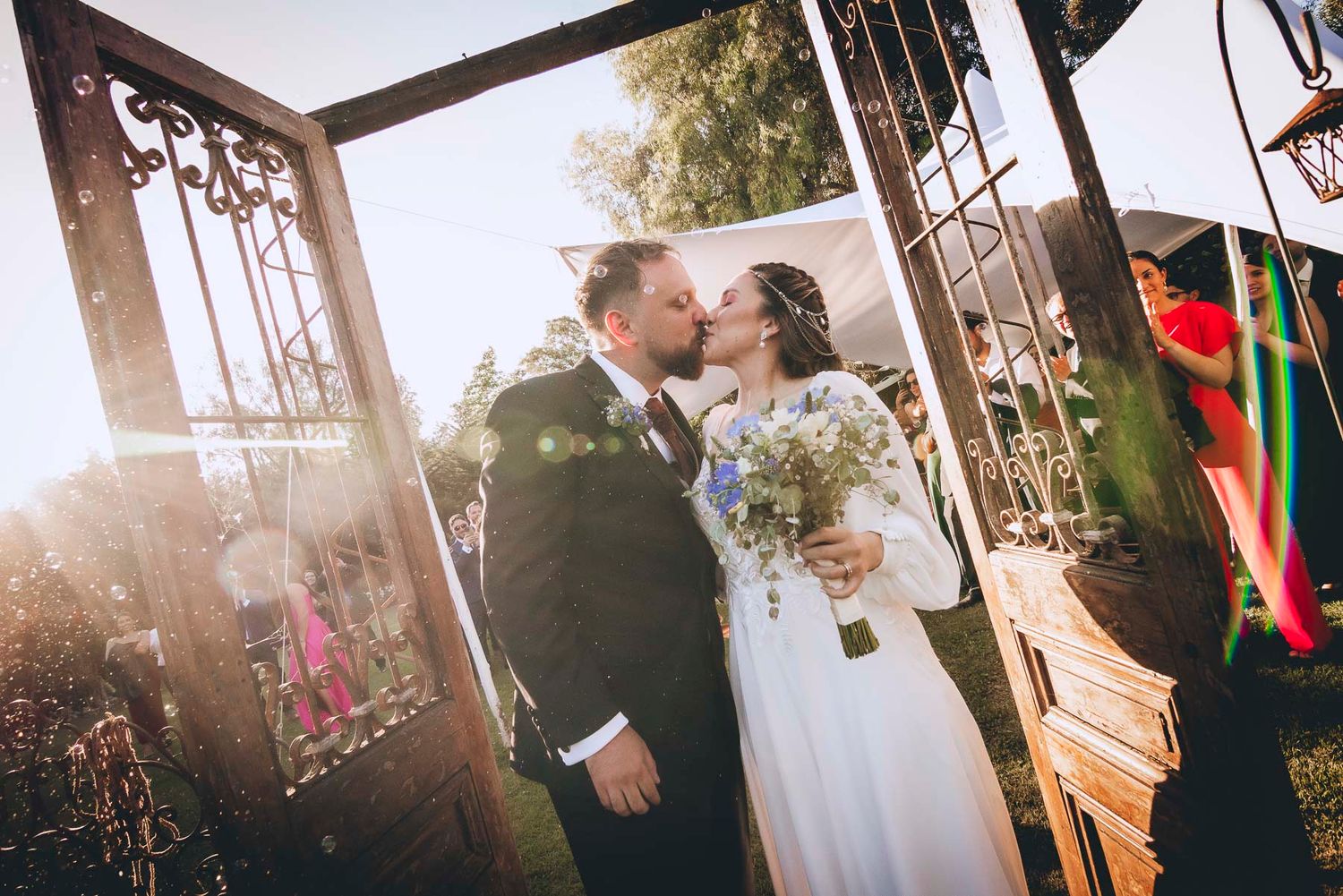 A wedding couple sharing a kiss under an ornate archway with sunlight streaming through.