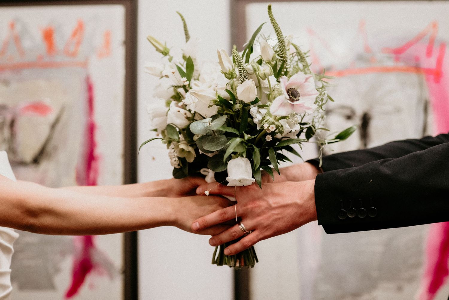 Two hands exchanging a delicate white and green floral wedding bouquet against a blurred background.