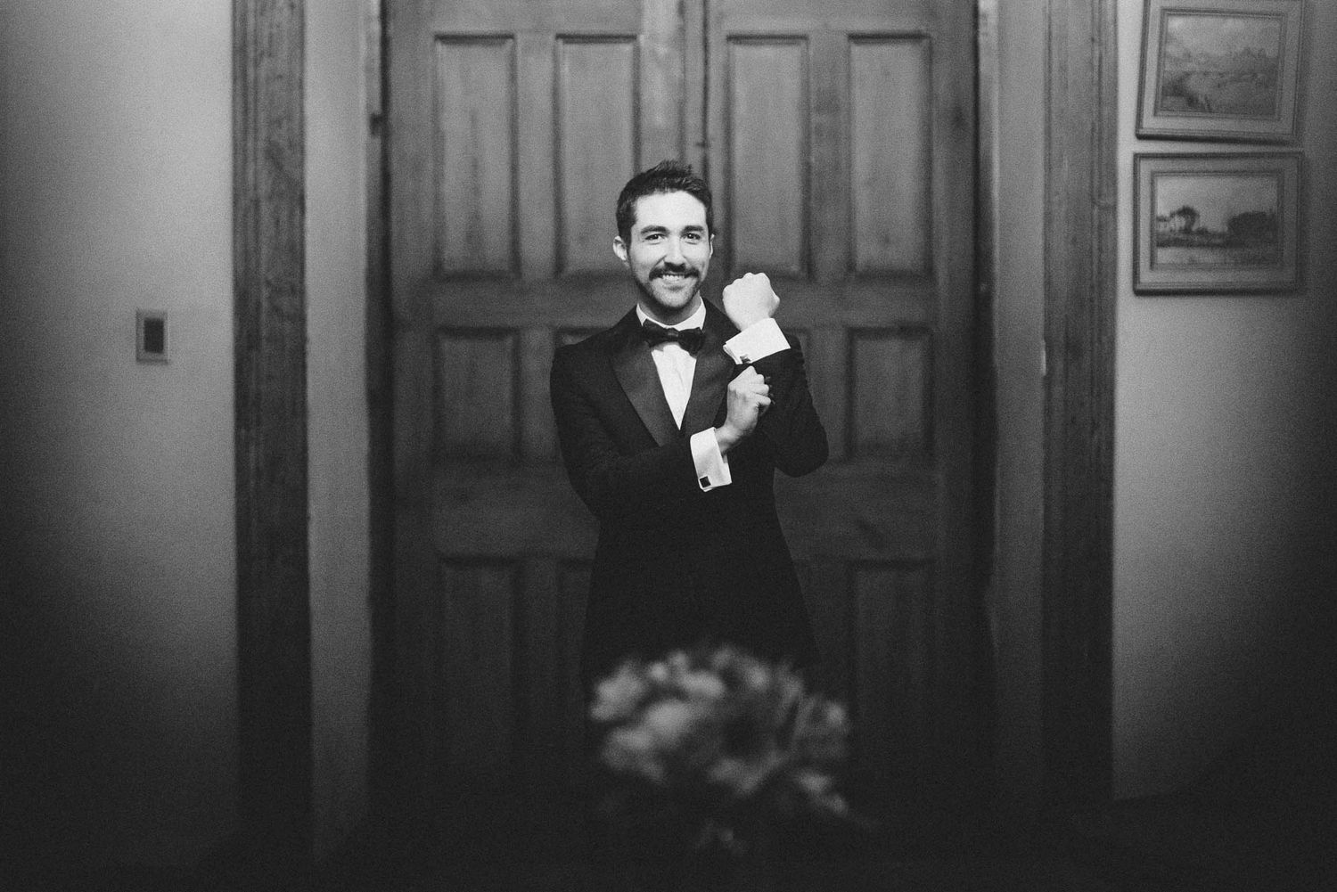 A black and white photo shows a groom in tuxedo holding a small white puppy in front of wooden doors.