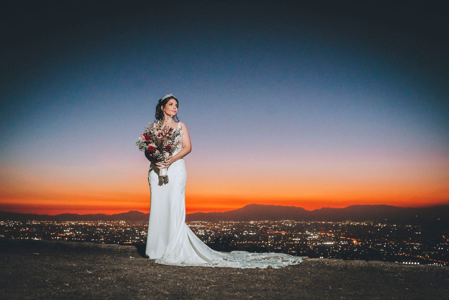 A figure in a flowing white wedding dress poses against a dramatic sunset cityscape.