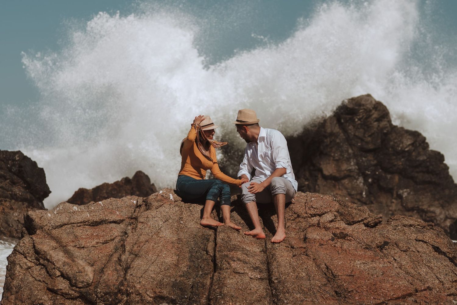Ocean waves crash against rocky coastline while people sit on cliffs overlooking the sea.