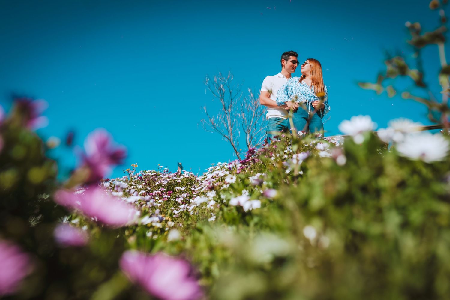 Romantic couple photographed among blooming flowers against bright blue sky.
