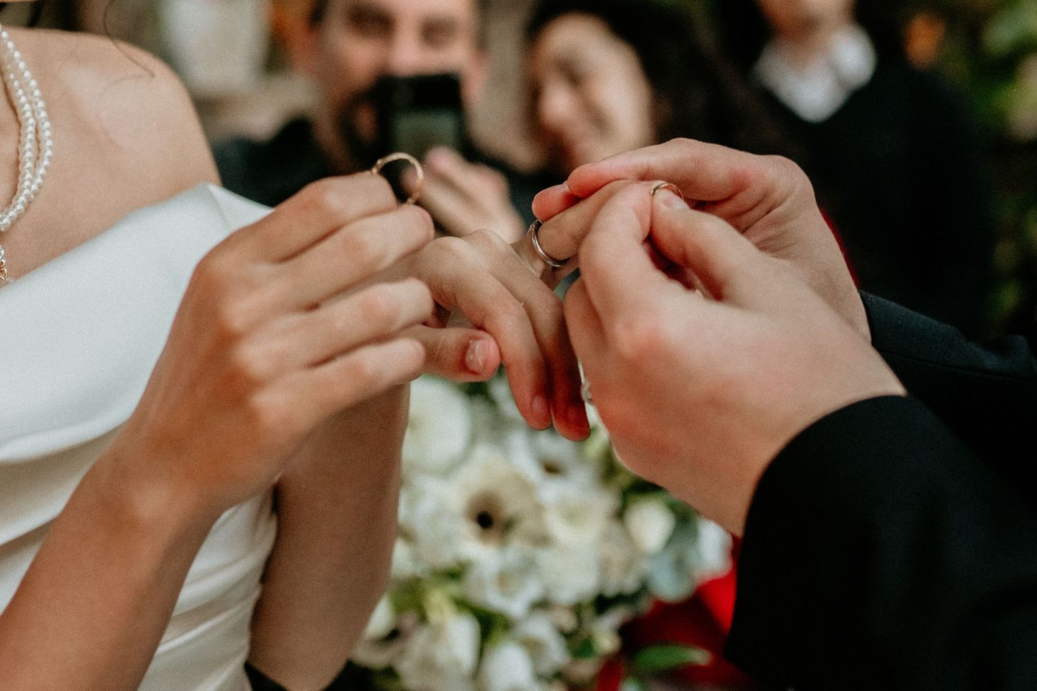 Close up of hands exchanging wedding rings during marriage ceremony with white floral bouquet in background.