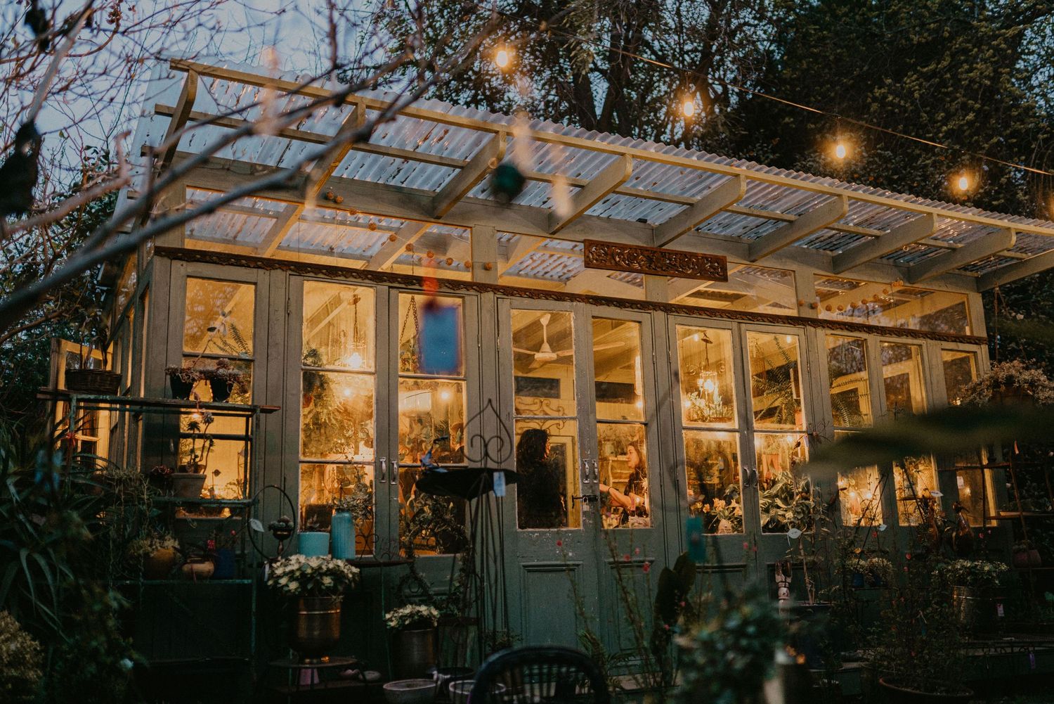 A rustic outdoor greenhouse restaurant glows warmly at dusk with string lights illuminating its glass and wood structure.