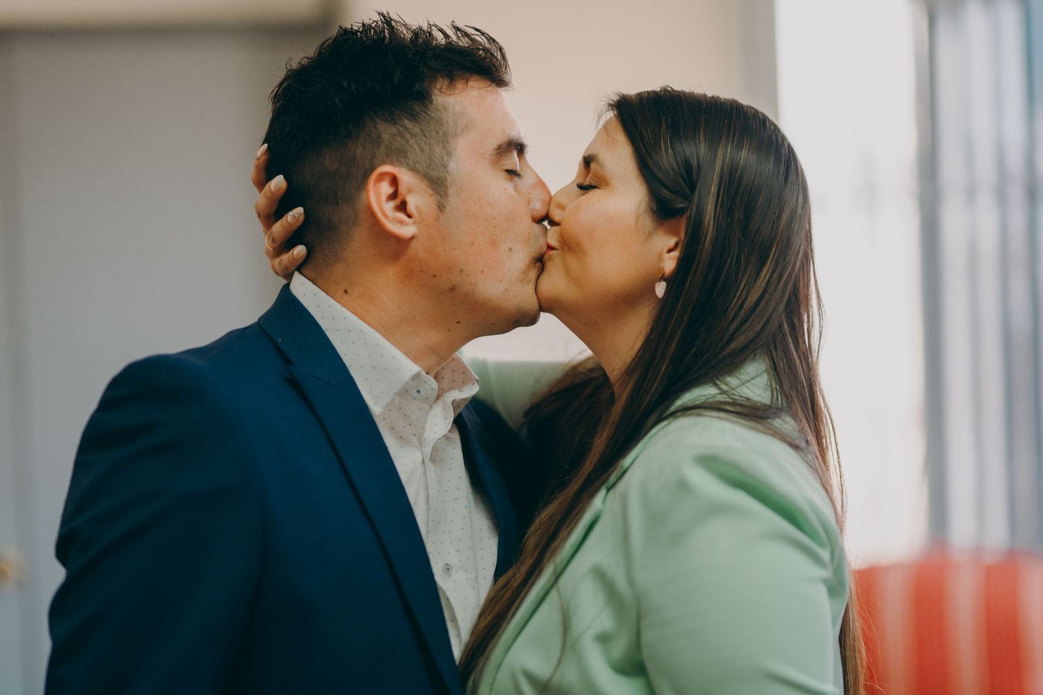A couple shares a romantic kiss by a window in a modern office setting.