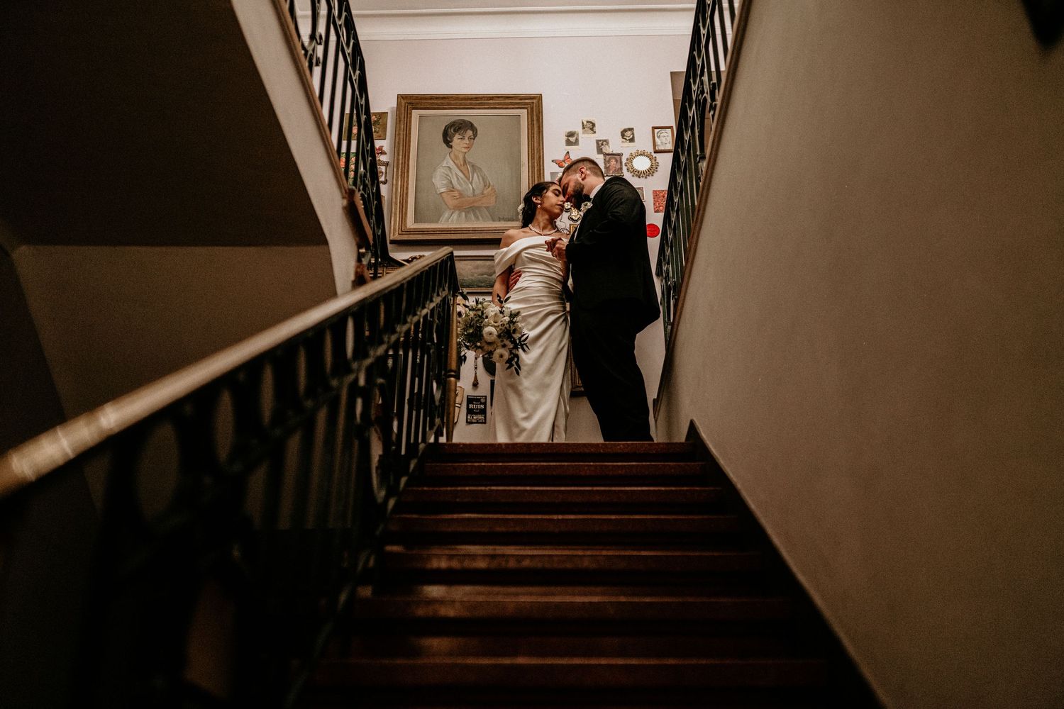A couple in wedding attire walks down a dark wooden staircase with a vintage framed photograph on the wall.