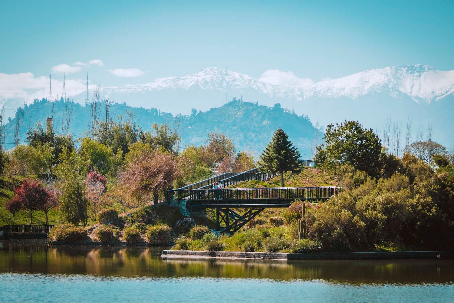 A scenic bridge spans across a lake with snow-capped mountains and blue skies in the background.