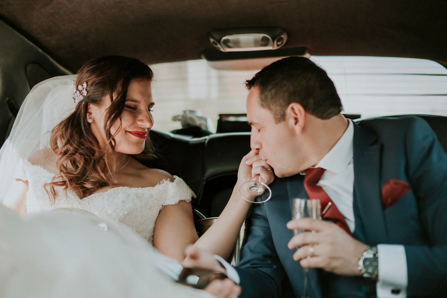 A romantic moment between newlyweds sharing champagne in the backseat of a car after their wedding ceremony.