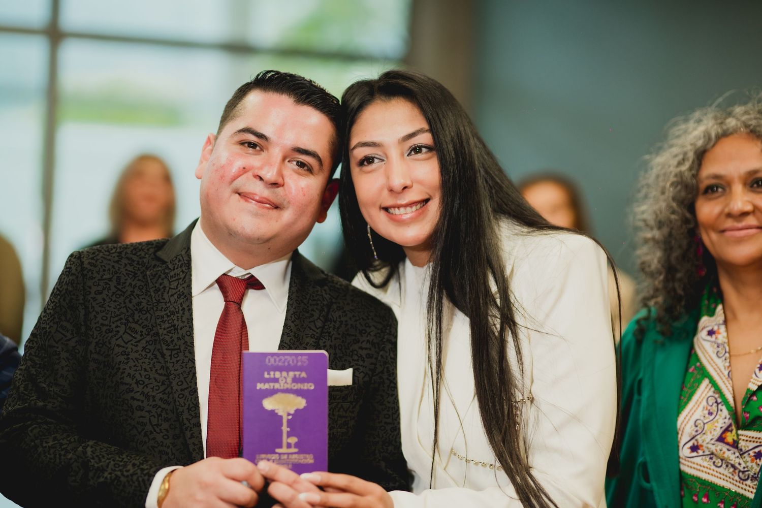 Two people smile at a formal event while holding a small purple book, with another person visible in the background.