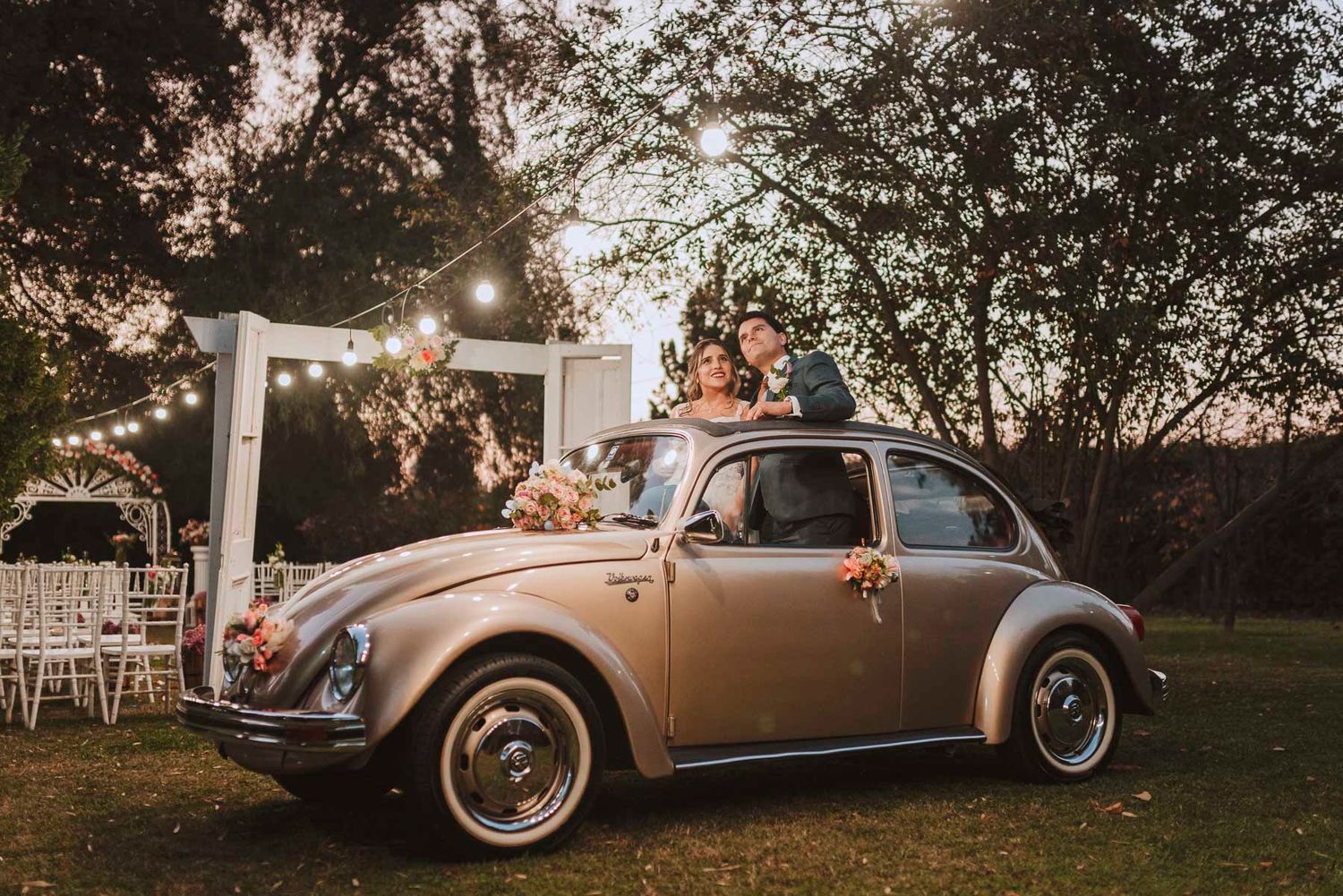 A vintage Volkswagen Beetle decorated with flowers at an outdoor evening celebration with string lights.