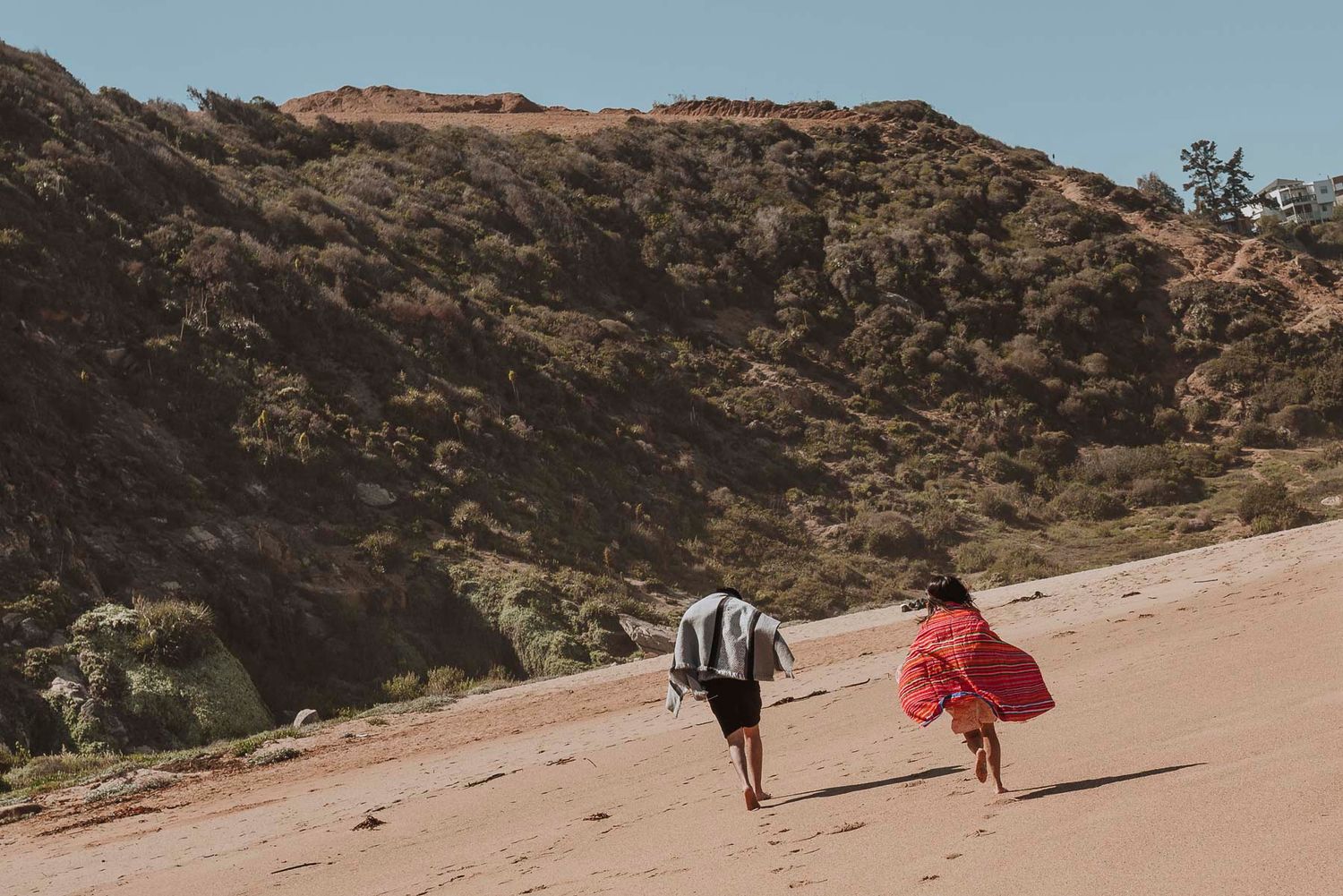 Two people run together on a sandy hill against a mountainous backdrop on a sunny day.
