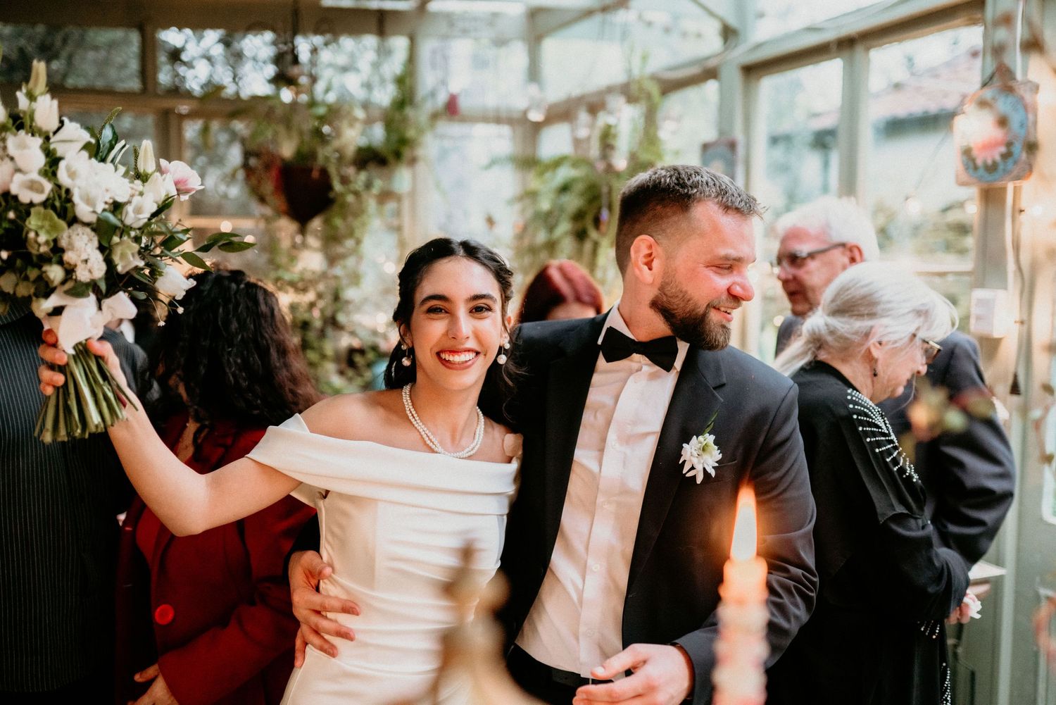 Happy couple celebrates during their wedding ceremony in a light-filled greenhouse setting with white floral decorations.