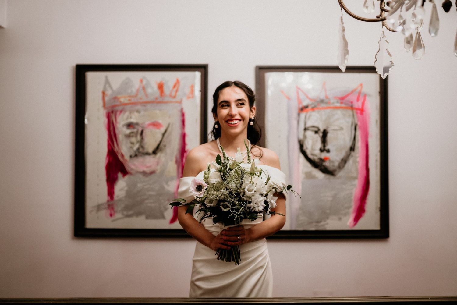 A person in a white wedding dress holds a stunning bouquet while standing in front of framed artwork.