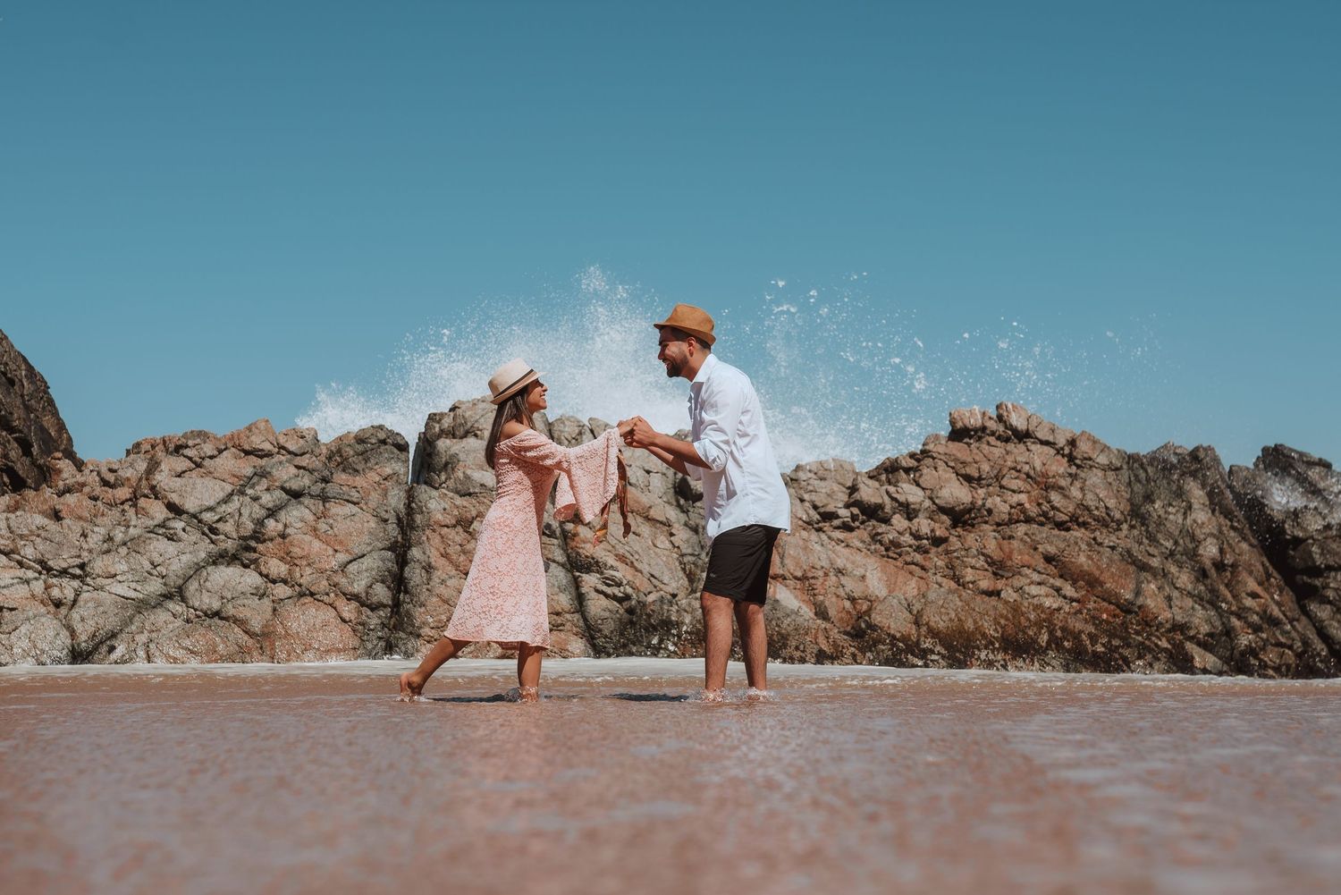 People enjoying sunny day at beach with waves splashing against rocky coastline.