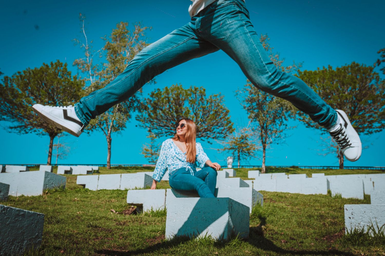 Person leaping playfully over someone sitting by rows of white headstones against a vibrant blue sky.