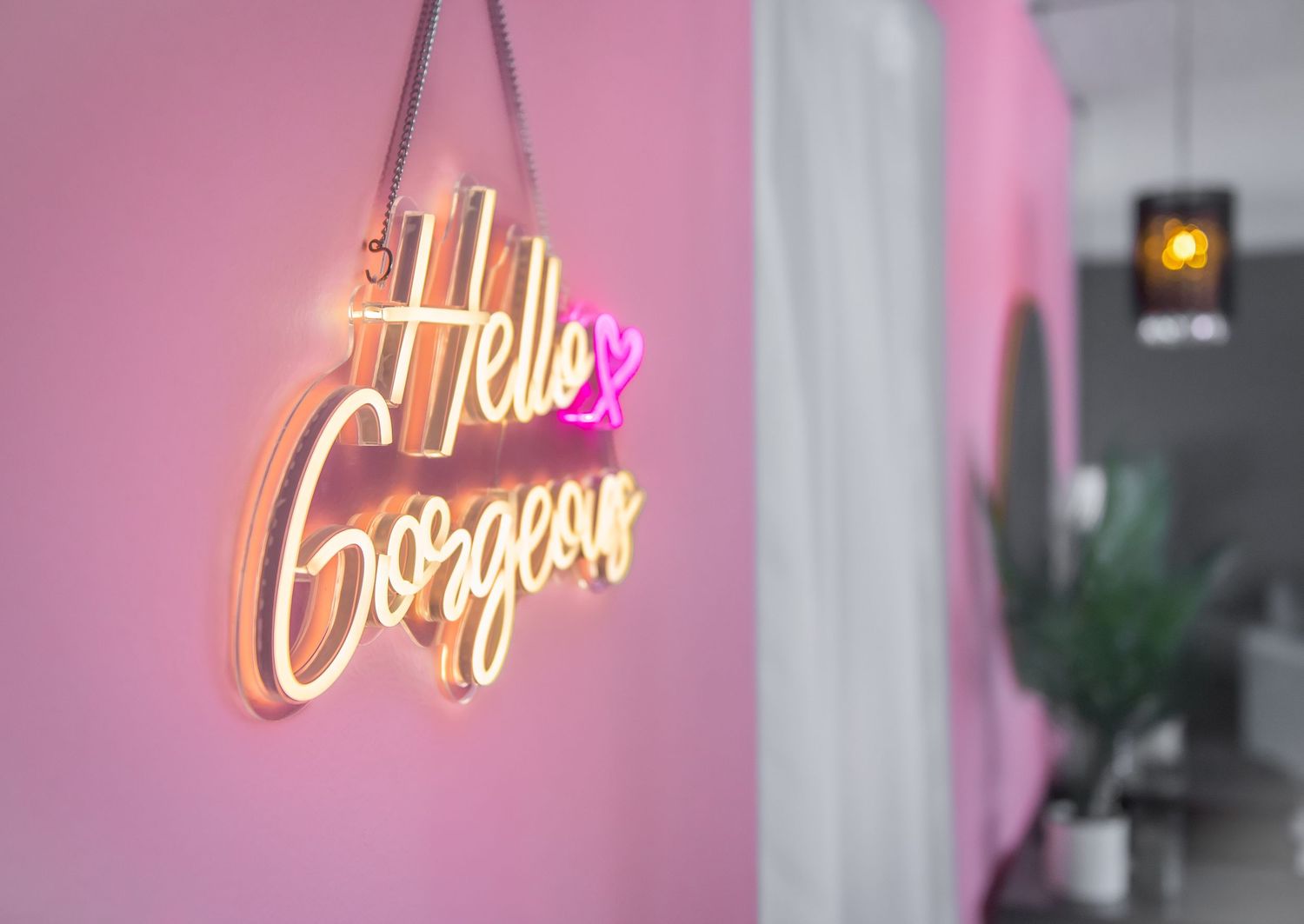 A neon sign reading 'Hello Gorgeous' glows against a pink wall with a green plant visible in the background.