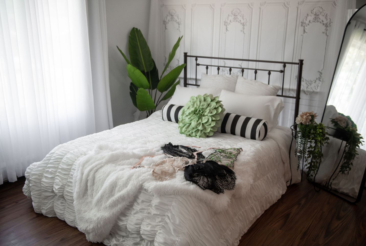 A cozy bedroom set featuring a white wrought iron bed with plush bedding, potted plants, and natural light streaming in.