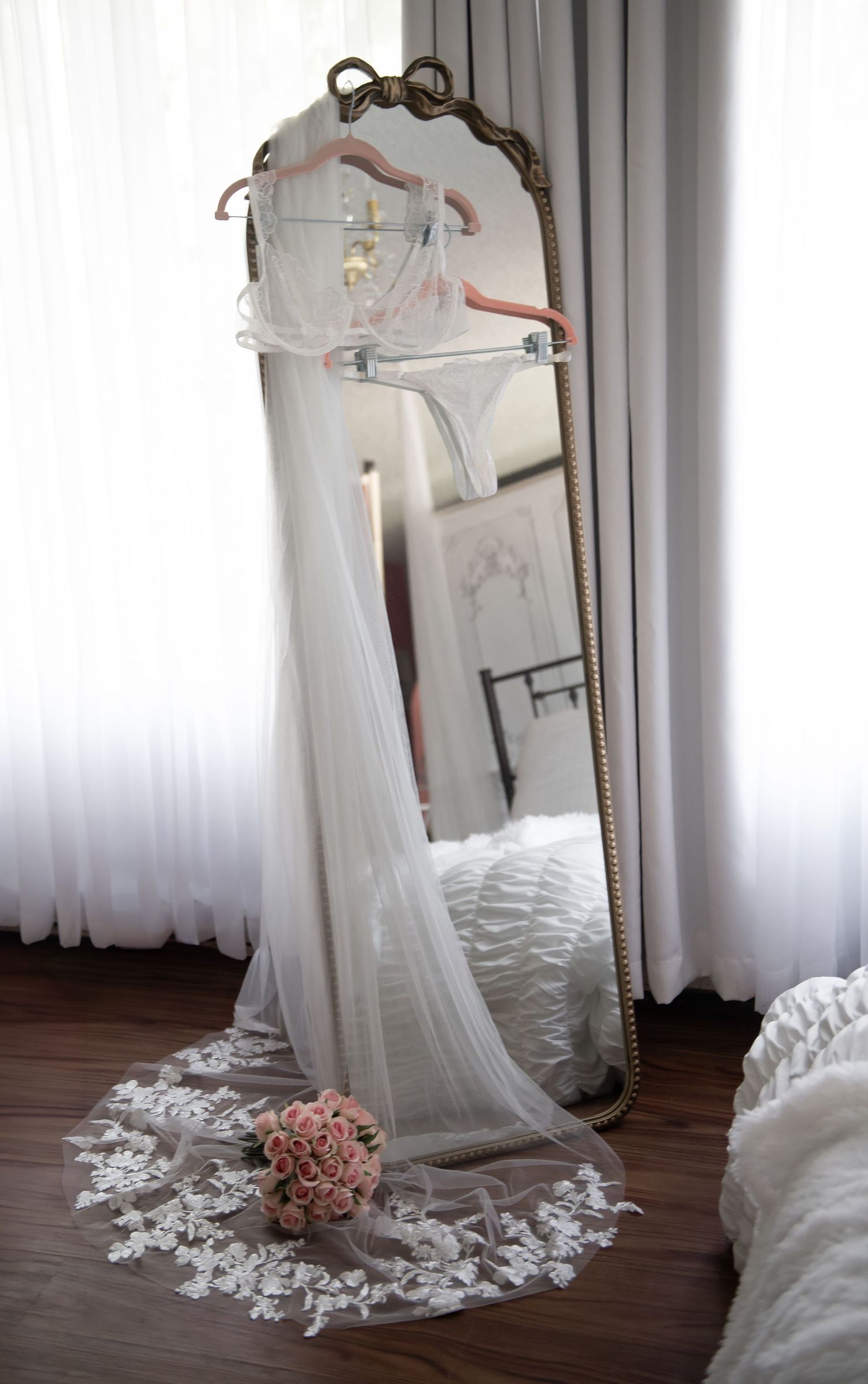A white wedding dress and veil displayed on hangers against a vintage mirror with lace details and blush pink accents.
