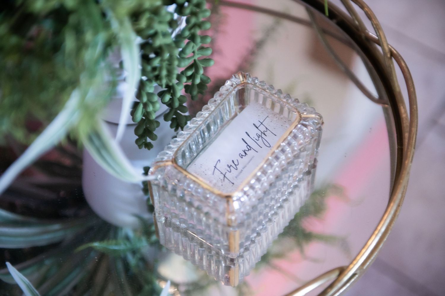 A vintage glass trinket box with decorative details sits on a mirrored surface near greenery and floral elements.