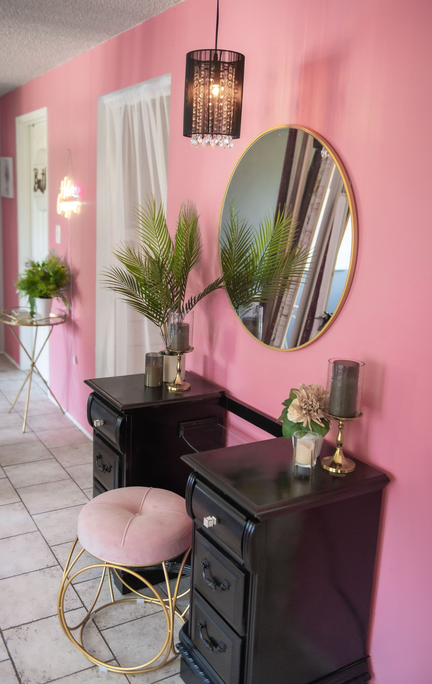 Modern vanity area with pink walls, round gold mirror, black dressers, palm plants and a chic pendant light fixture.