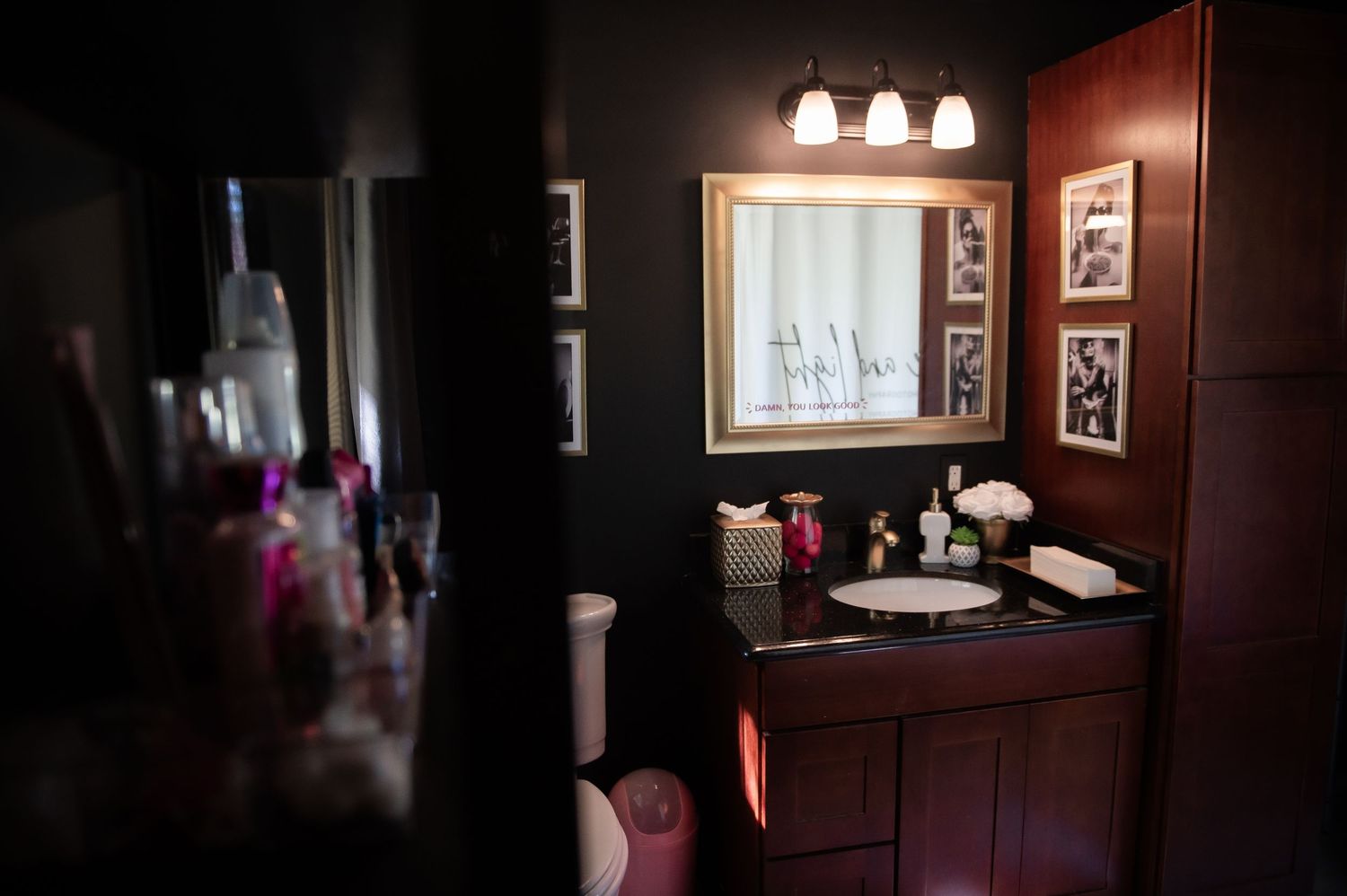 A dimly lit bathroom with black walls features framed artwork and warm lighting creating an intimate gallery atmosphere.