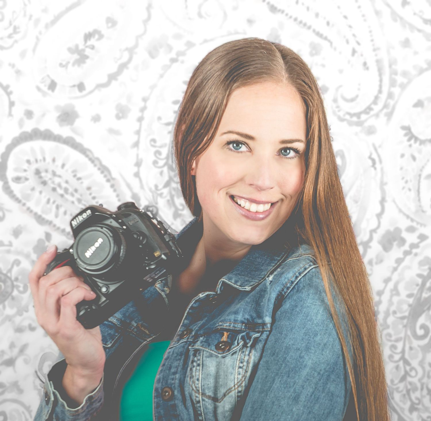 A boudoir  photographer wearing a denim jacket holds a professional DSLR camera against a decorative paisley background.