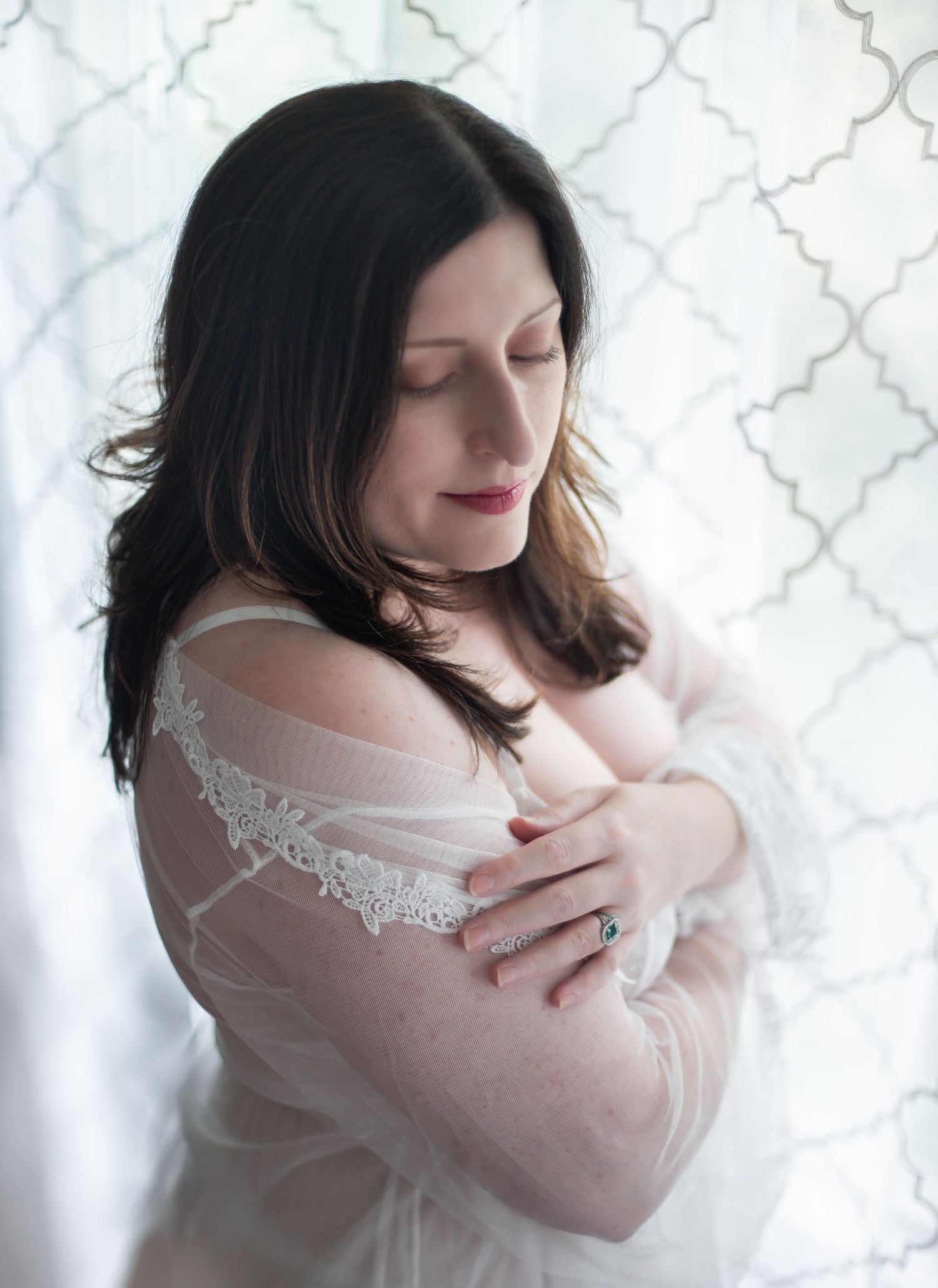 A woman in a delicate white lace robe poses against a patterned background in soft natural light.