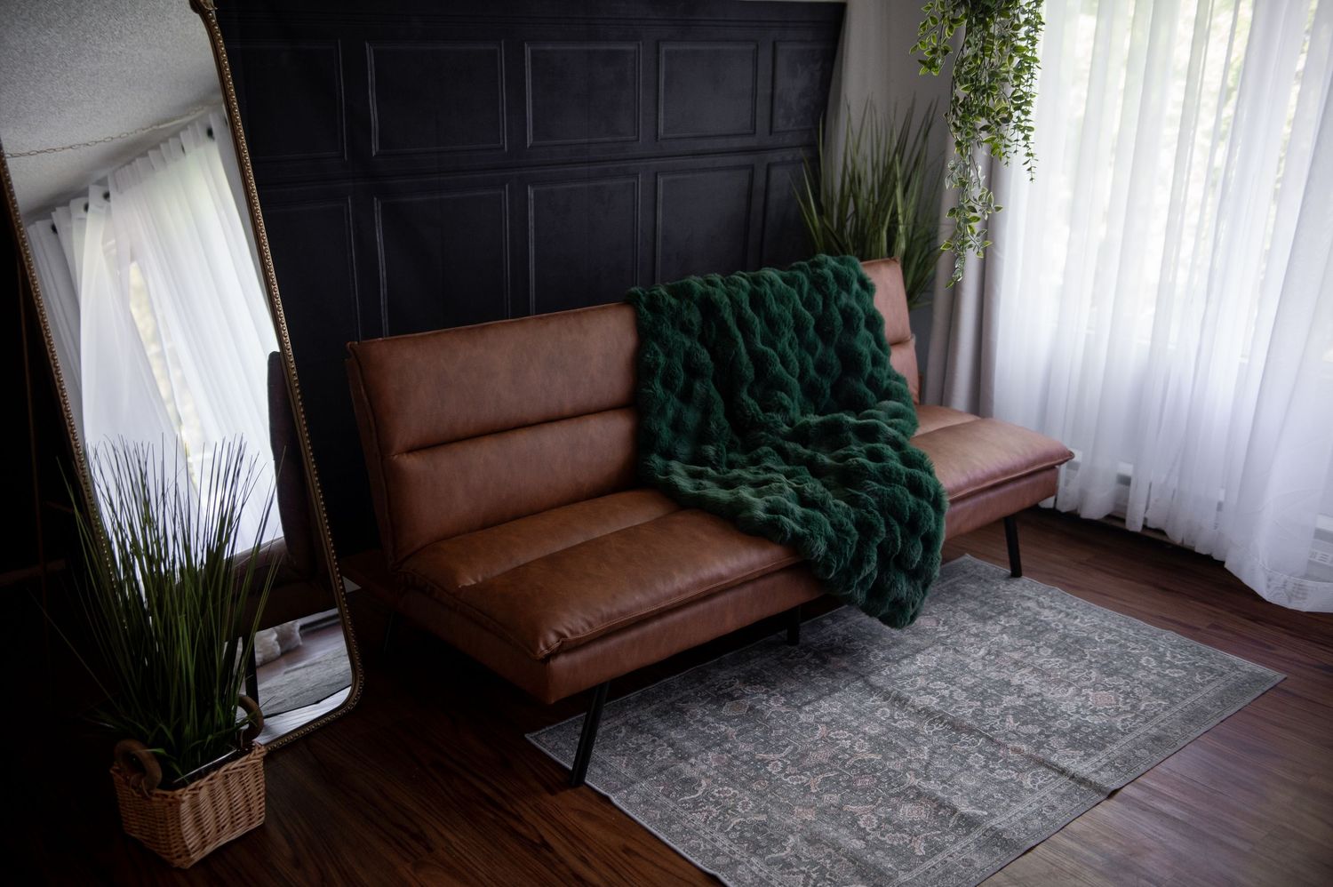 Brown leather mid-century modern sofa with emerald green throw blanket on hardwood floors beside sheer curtains.