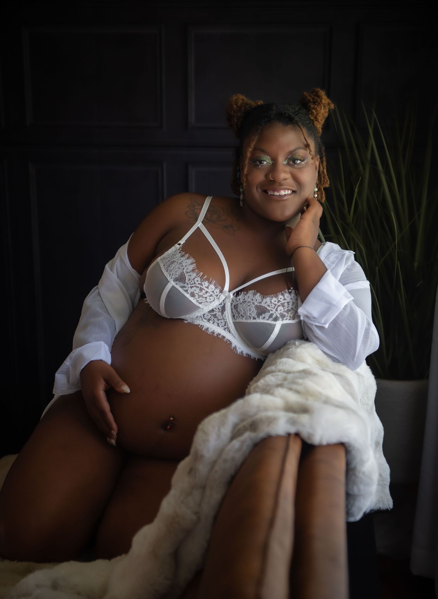 A pregnant woman wearing white lingerie and an open shirt poses on a couch with a blanket in a dimly lit room.