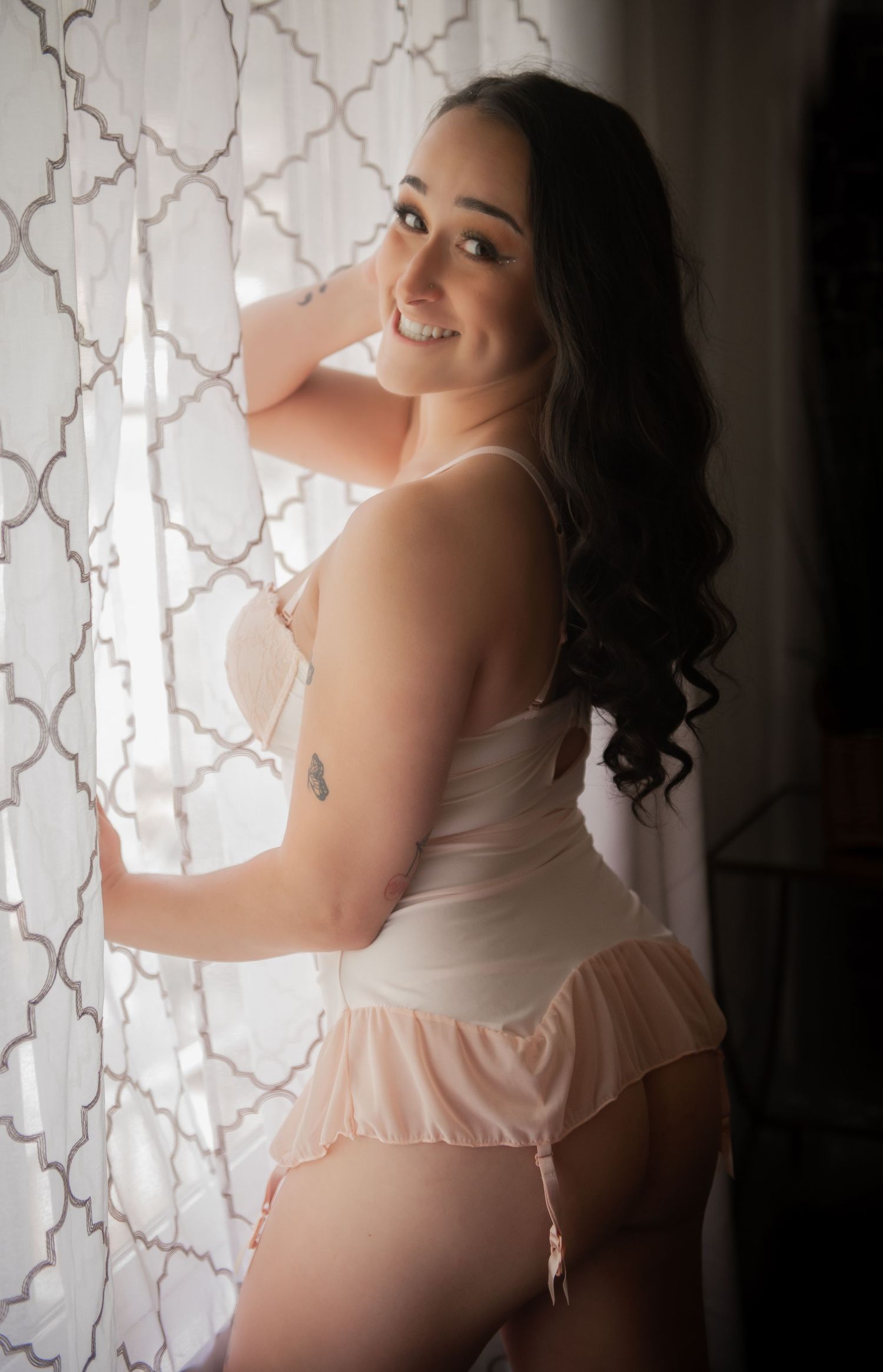 Soft backlit boudoir portrait by window with lace curtains in white lingerie.