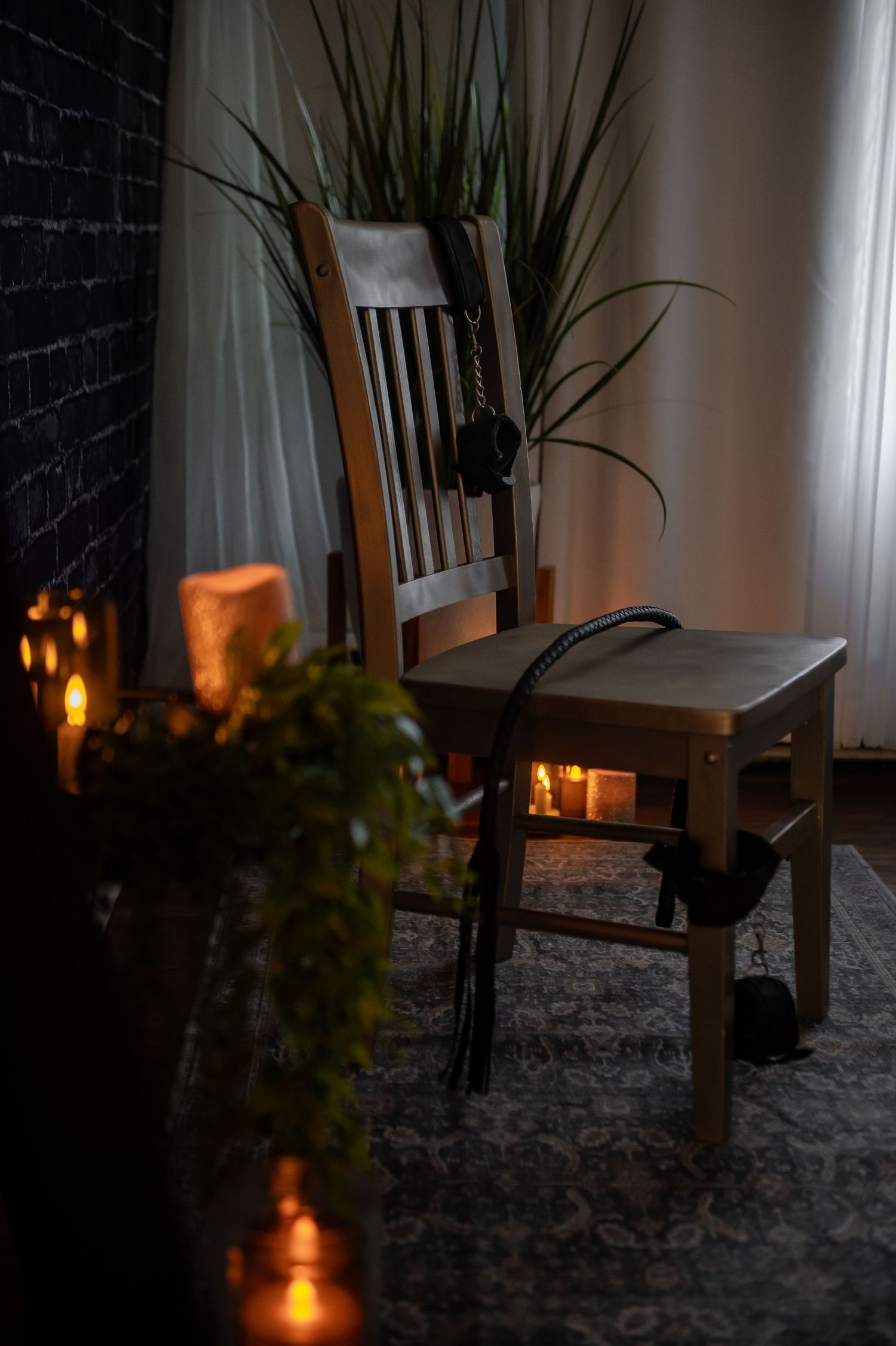 A wooden chair sits in moody lighting with glowing candles and plants creating a cozy atmospheric scene for naughty boudoir shots.