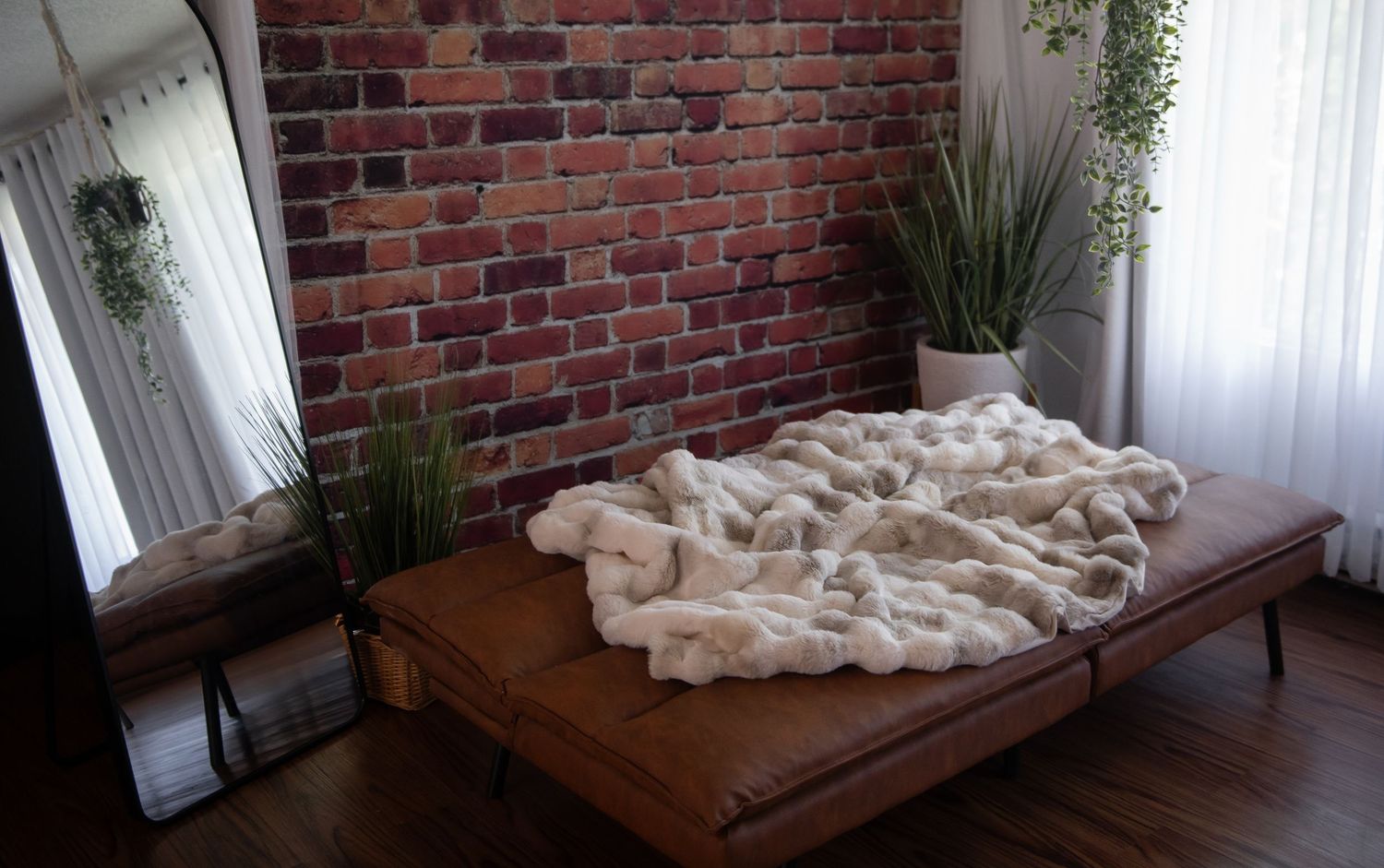 A rustic bedroom set with exposed brick wall features a brown chaise lounge with rumpled fur blanket and potted plants.