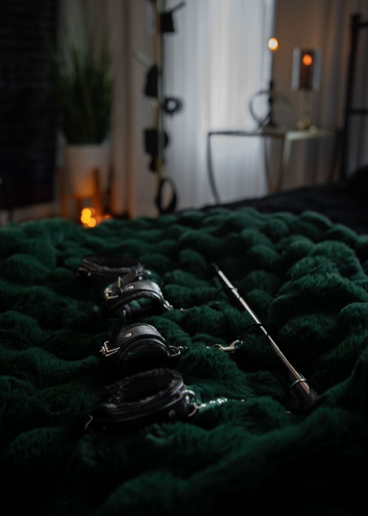 Dark leather spreader bar resting on a plush emerald green fur blanket with soft ambient lighting in naughty boudoir set.