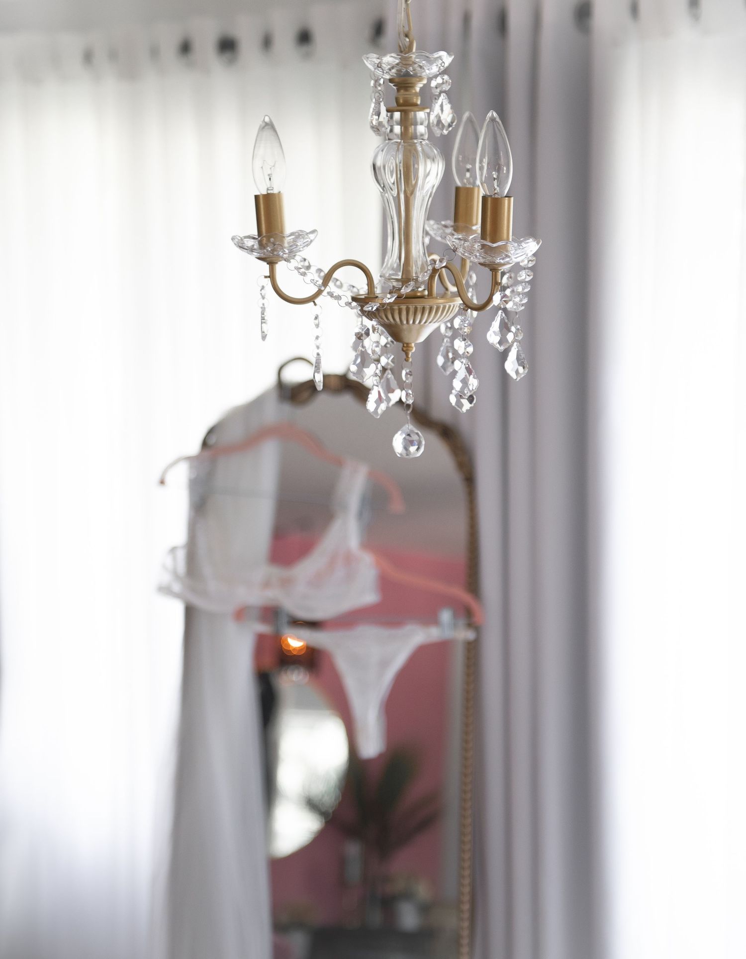 Crystal chandelier with draped bridal lingerie and candle-style lights hangs against white textured curtains in an elegant boudoir setting.