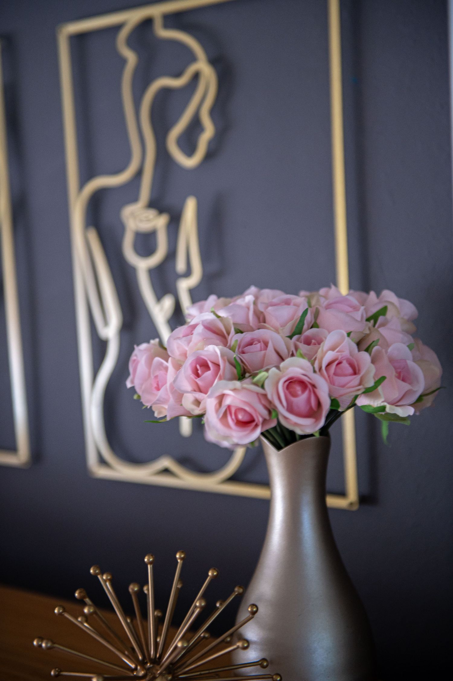 Pink roses in a matte gray vase sit beside decorative wall art featuring a linear figure design on a dark background.