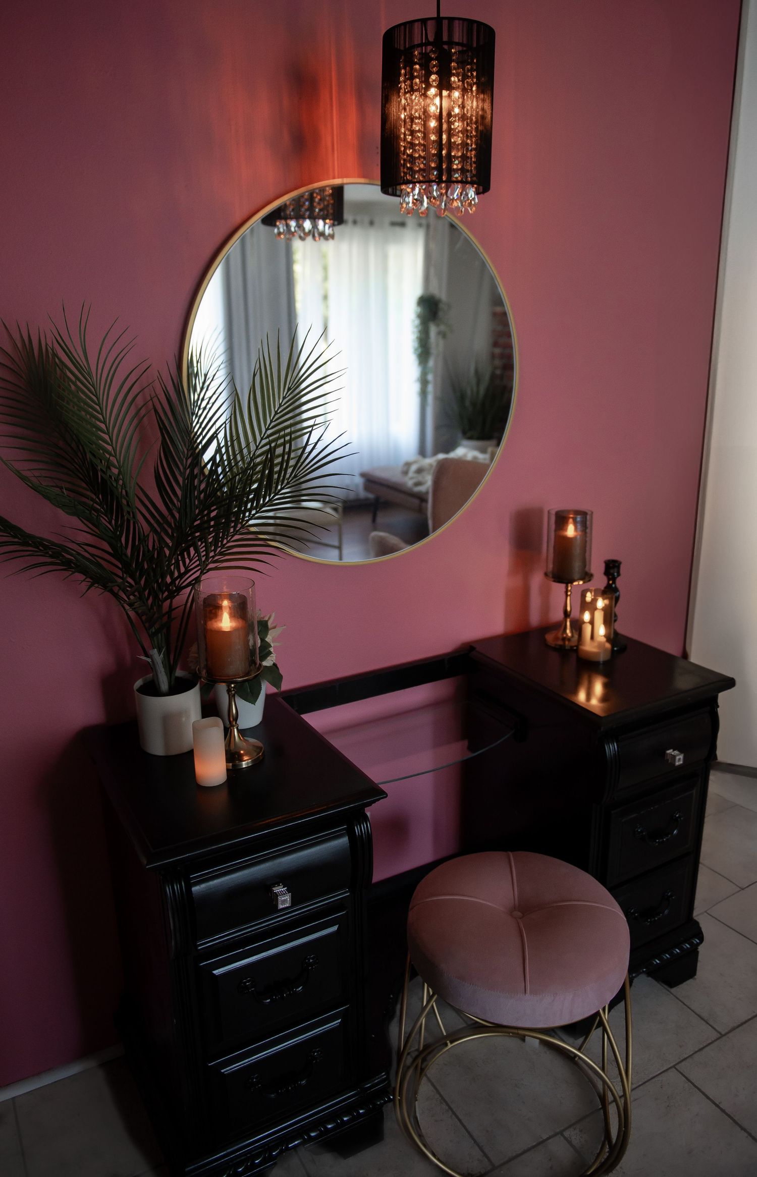A modern vanity setup with a round mirror, black furniture, potted palm plant, and hanging crystal lamp against a pink wall.