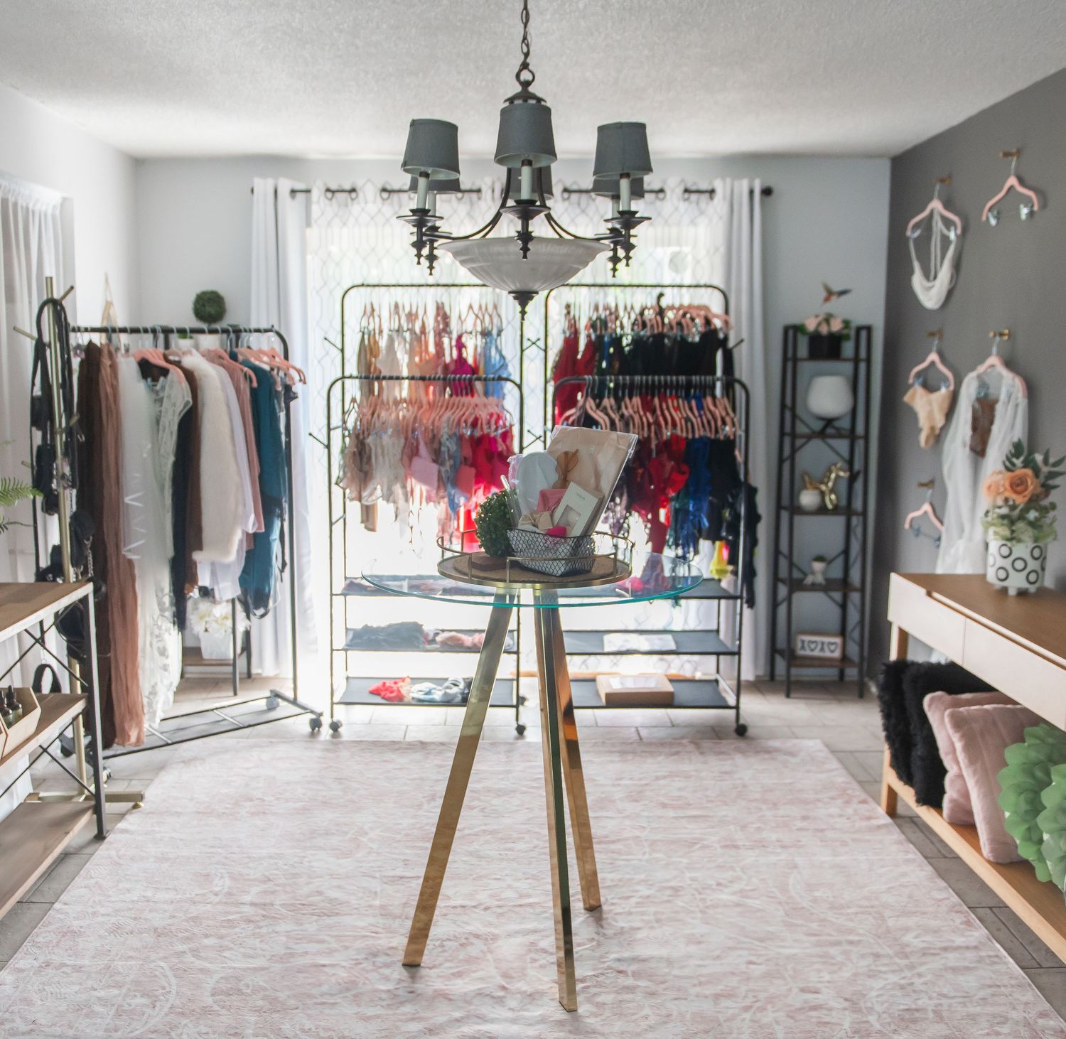 A vintage-style boutique interior with clothing racks, display shelves, and an elegant chandelier in a bright, organized boudoir space.