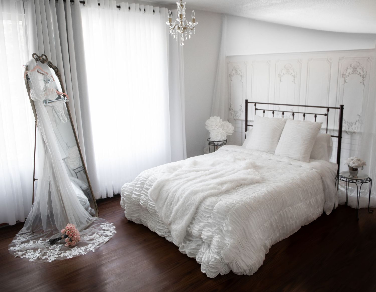 Elegant white bedroom with lace bedding, vintage floor mirror, crystal chandelier, and flowing white curtains on dark hardwood floors.
