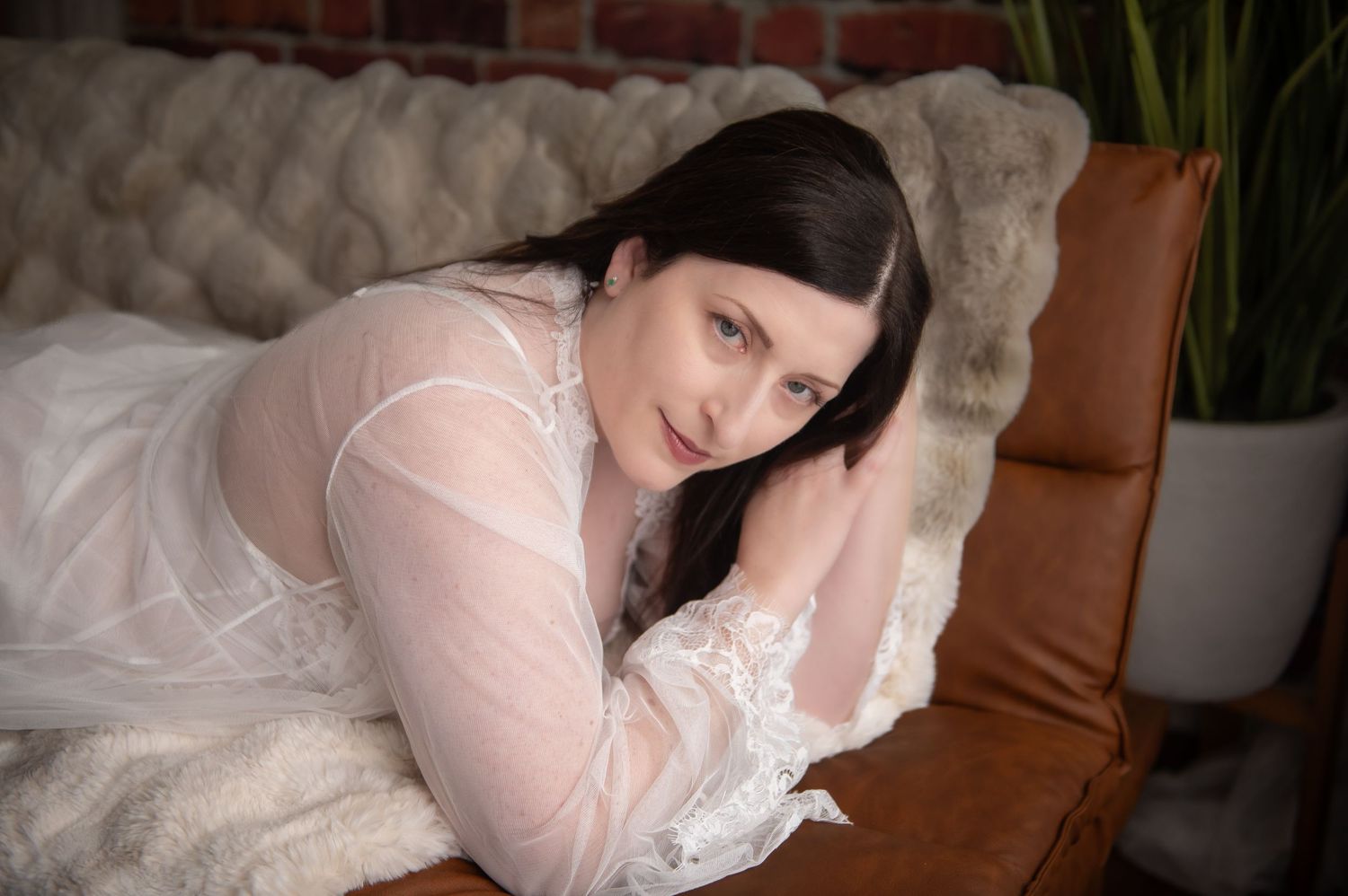 A person in a white lace gown lies on a couch with brown pillows, with indoor plants in the background.