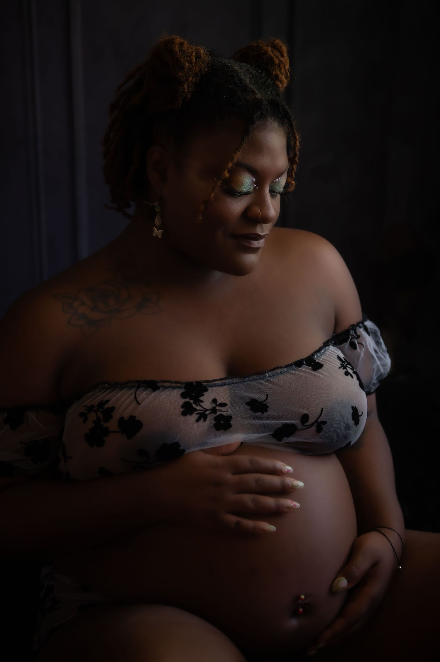 Intimate maternity portrait in moody lighting showing an off-shoulder floral lingerie piece and tender moment with belly.