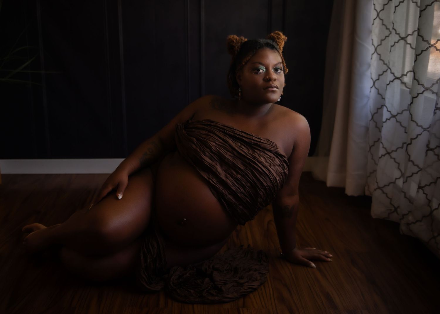 Dark, moody maternity boudoir photograph taken in dramatic lighting against decorative curtains.